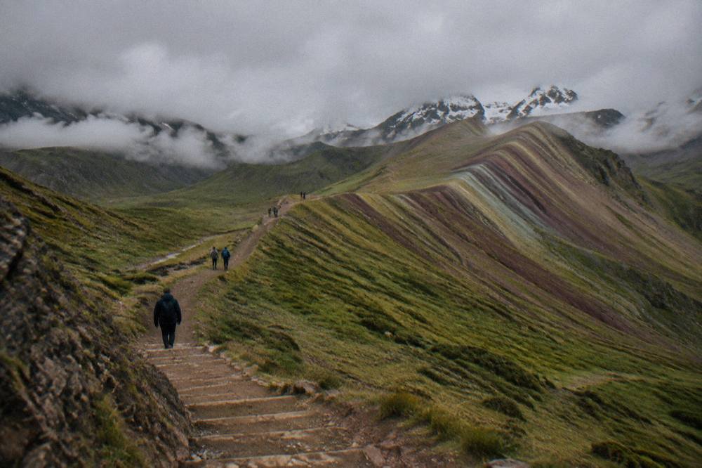 Walkers on a dirt path climbing a high ridge toward striped mineral slopes with snow on distant peaks