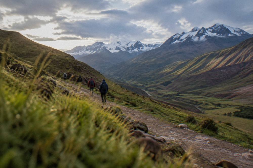 Walkers in warm jackets on a high dirt trail between green valley slopes and distant snow-capped peaks