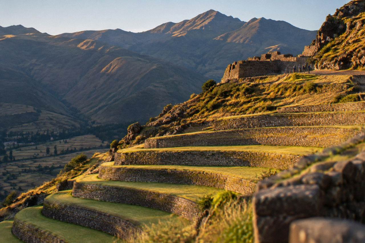 Terraced Inca agricultural steps on a green hillside in the Sacred Valley near Cusco, with mountains behind under a clear sky.