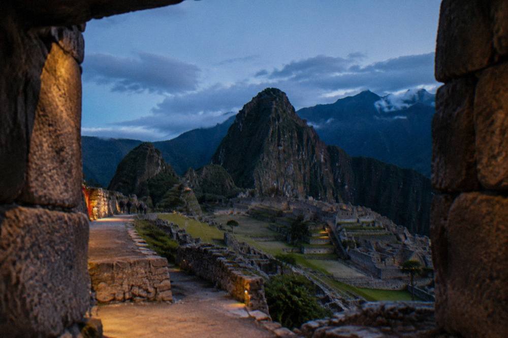 Stone terraces and ruins of Machu Picchu on a ridge with green peaks rising behind under scattered cloud.