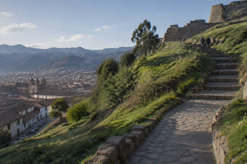 A paved walking path and stone steps climbing a green hillside above Cusco toward ancient walls