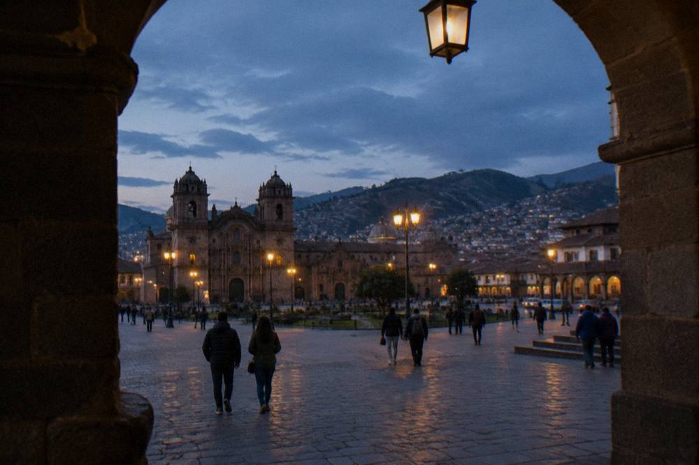 Visitors in warm layers and jackets crossing a stone plaza in Cusco at dusk, seen from behind