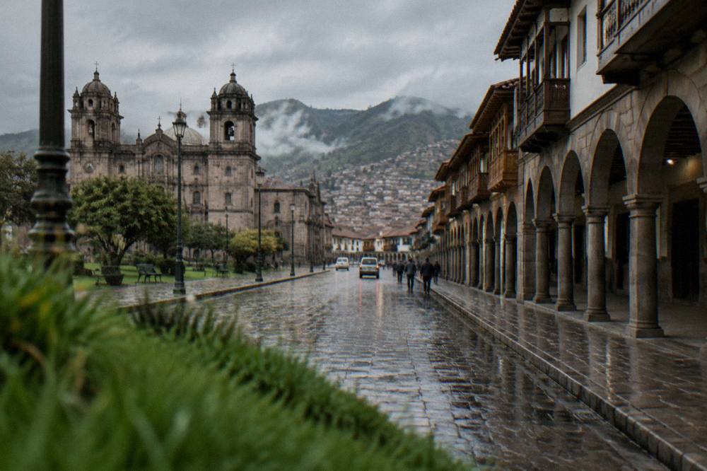 A quiet stone-paved street in Cusco under grey afternoon sky with wet flagstones reflecting the colonial arcades