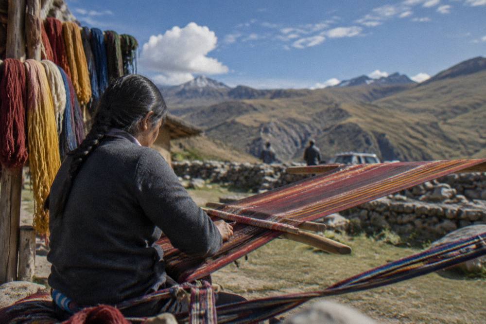 A weaver working threads on a traditional loom with coloured yarns hanging nearby