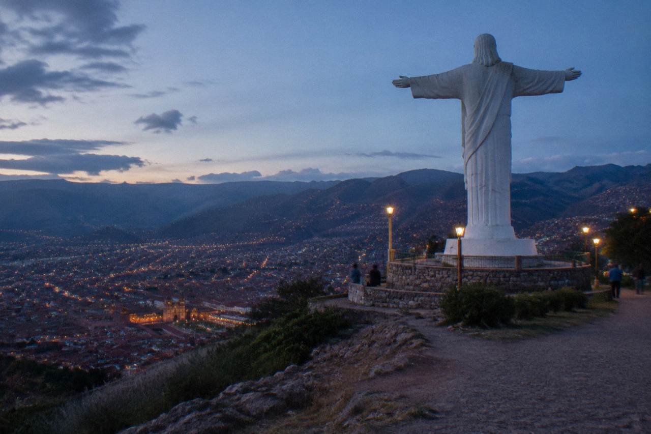 A tall white statue on a green hillside above the red rooftops of a high Andean city