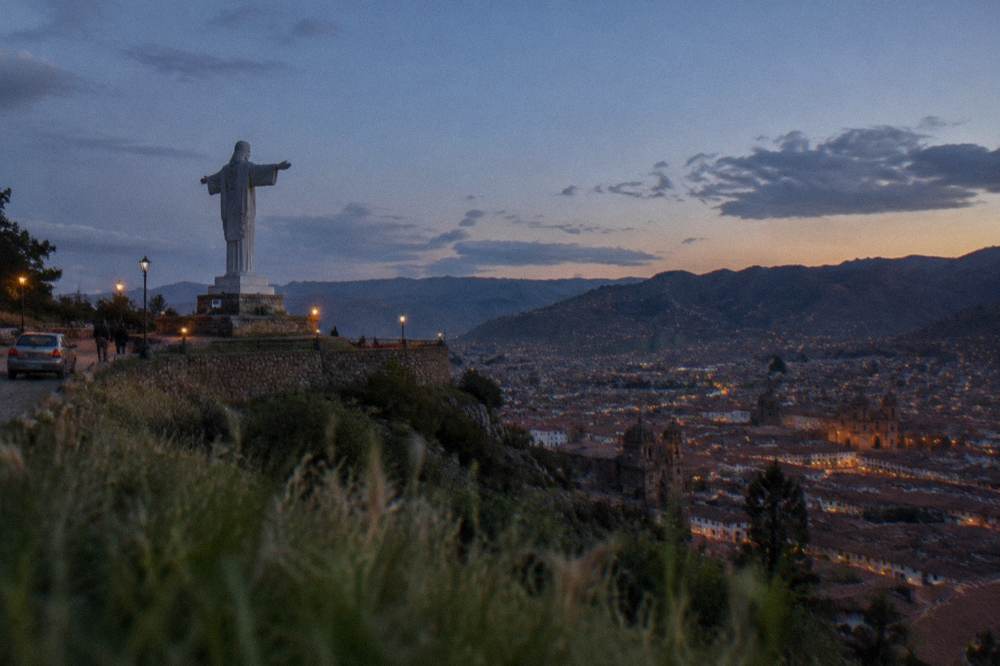 A white statue with open arms silhouetted against a wide sky above a highland city