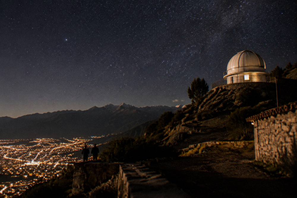 A telescope dome on a dark hillside under a starfilled sky above city lights
