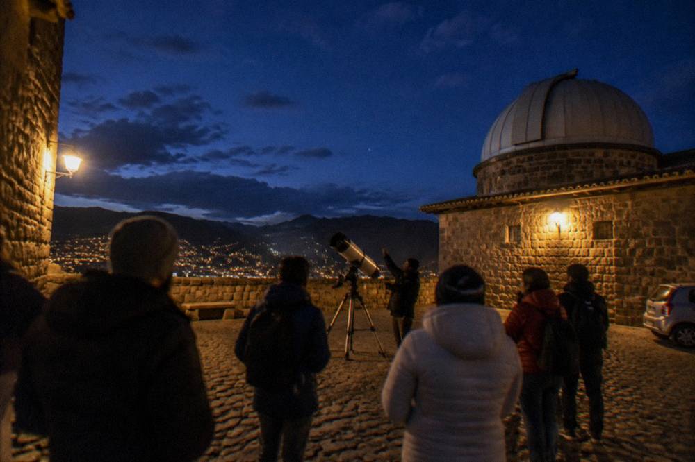 A group of visitors in warm coats listening to a guide beside a telescope at night
