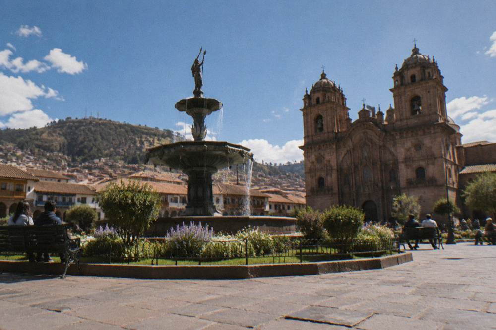 A stone fountain and gardens at the centre of the square with people resting on benches