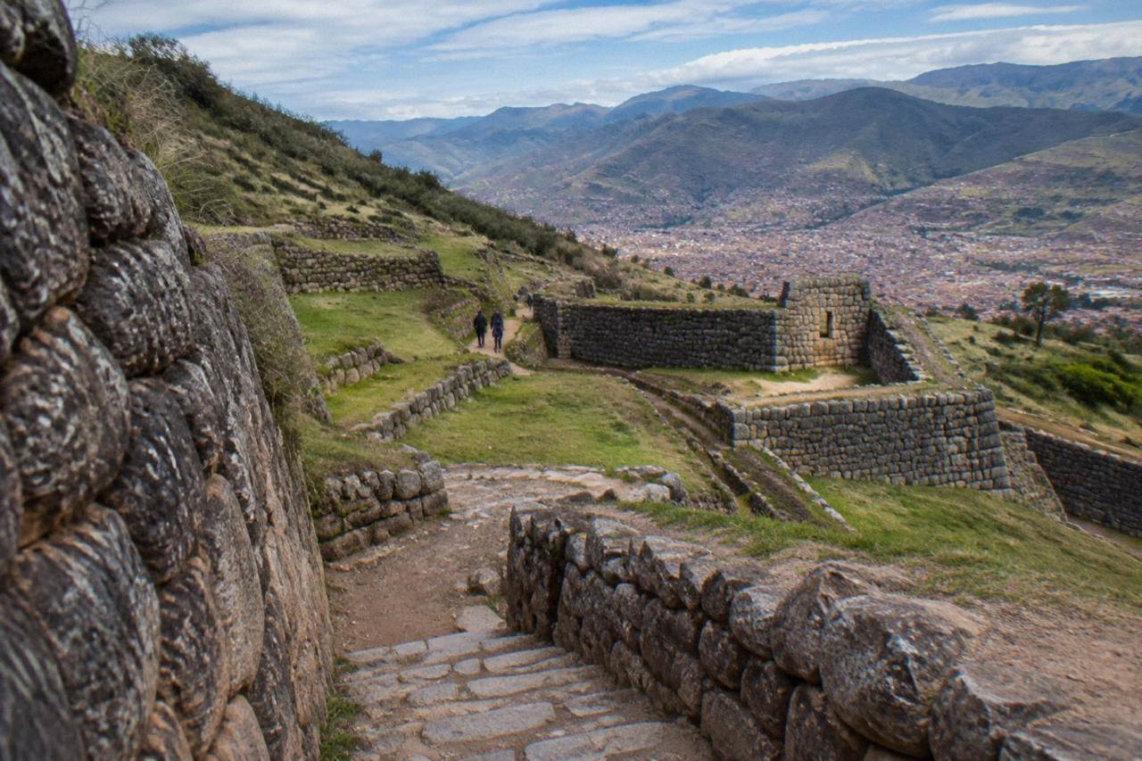 Stone terraces and walls of an Inca site on a green hillside above Cusco under a blue sky