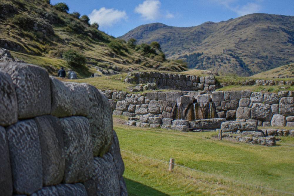 Rough stone walls and a grassy enclosure at an Inca site with hills in the background