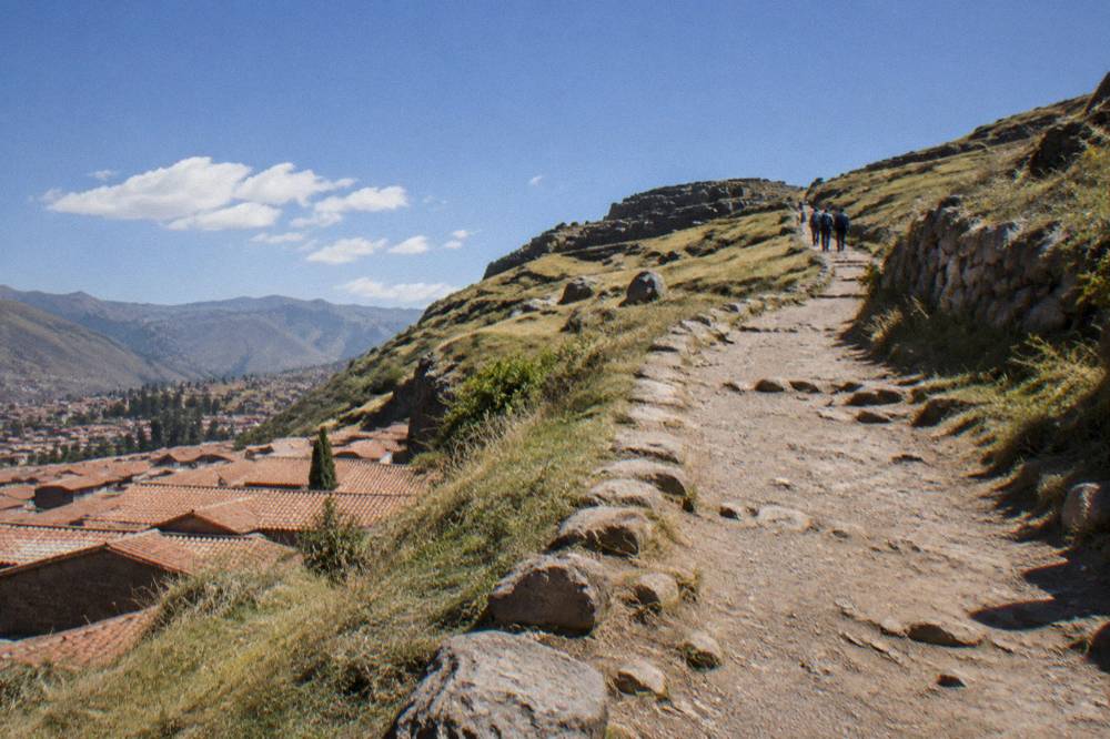 A hillside dirt path above red-tiled rooftops climbing toward open grassland