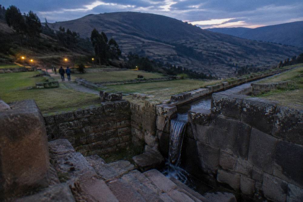 A stone aqueduct channel crossing a terrace wall at Tipon with hills behind