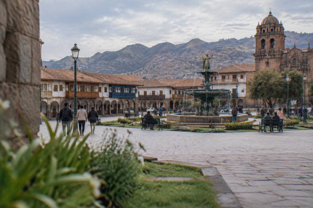 A sunlit stone square with colonial arcades and a fountain busy with people in dry season light