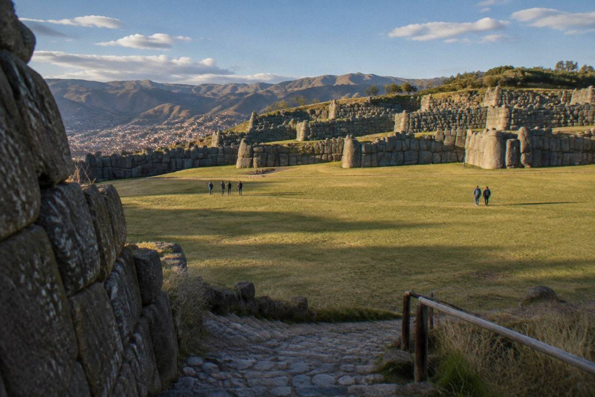 The terraced fitted-stone walls of Saqsaywaman across the grass esplanade under a blue sky, a few small visitors far off