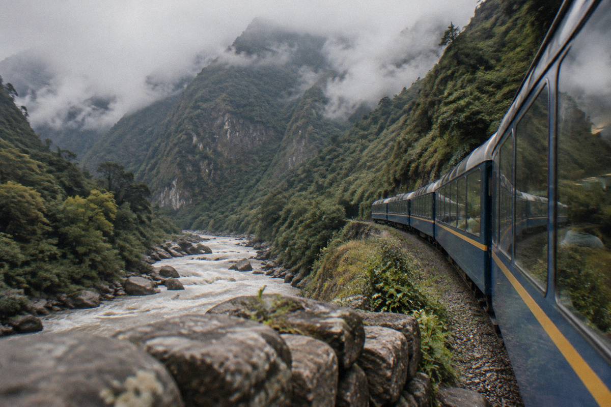 A passenger train with large panoramic windows running along a river gorge below steep forested mountains near Aguas Calientes