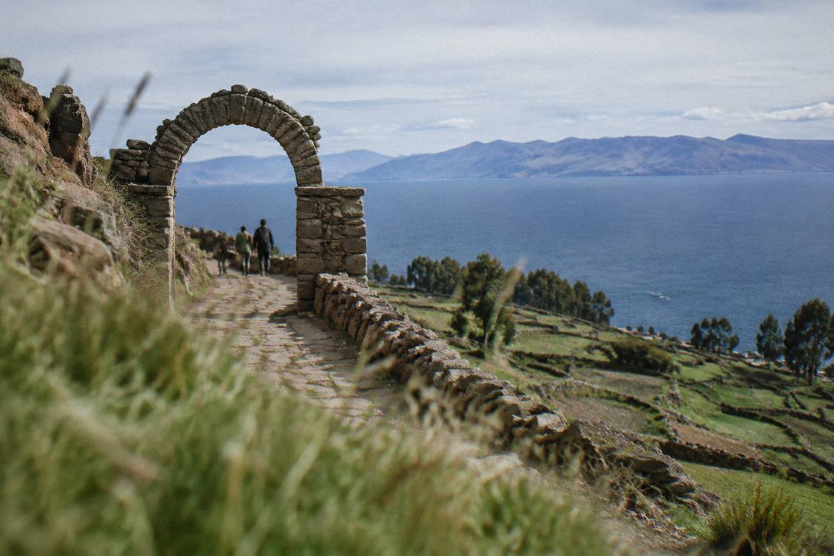 A stone arch on a hill path on Taquile Island above deep blue Lake Titicaca, terraced fields and the far shore beyond