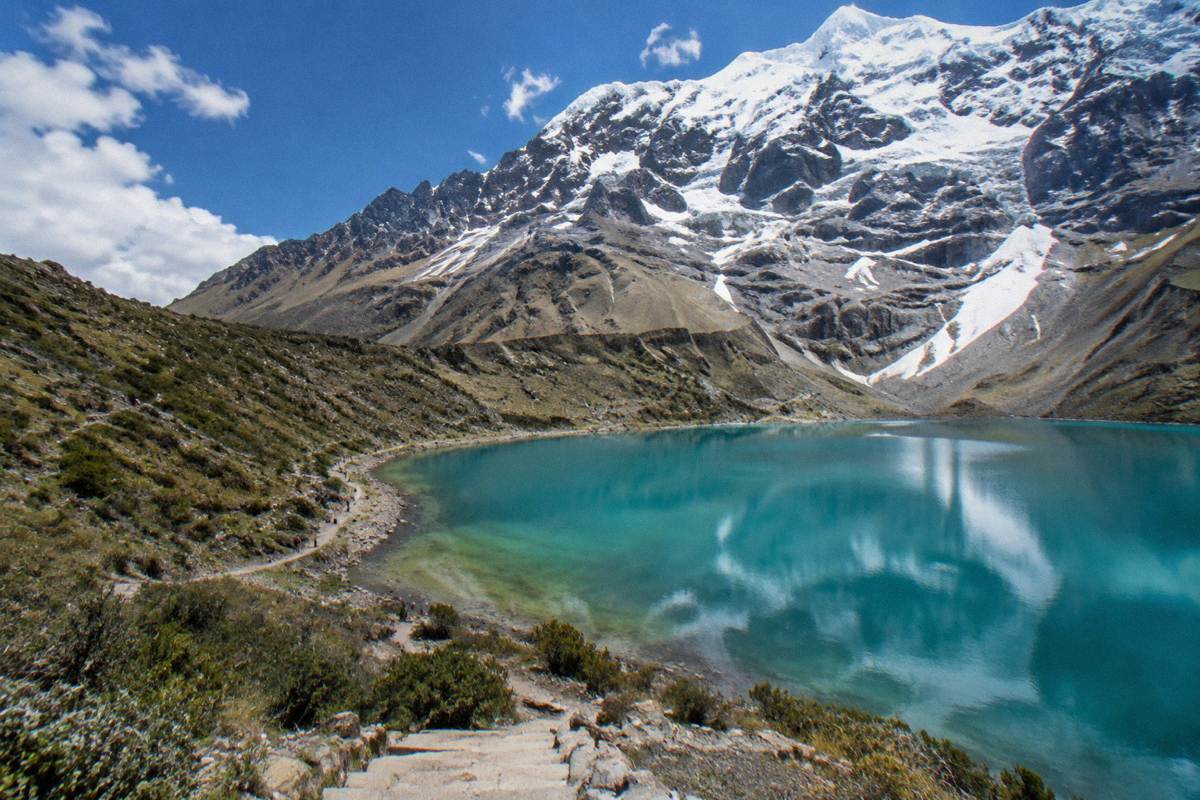 The turquoise glacial lake Humantay at the foot of a steep snowy peak, a narrow shoreline path with a few tiny distant walkers