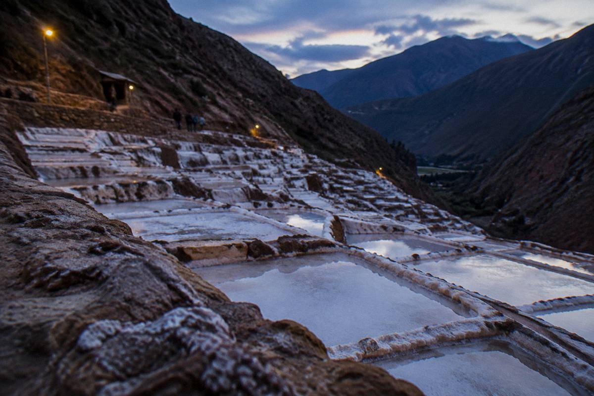 Terraced white salt pools stepping down a steep valley side in afternoon light, narrow earthen walkways between them