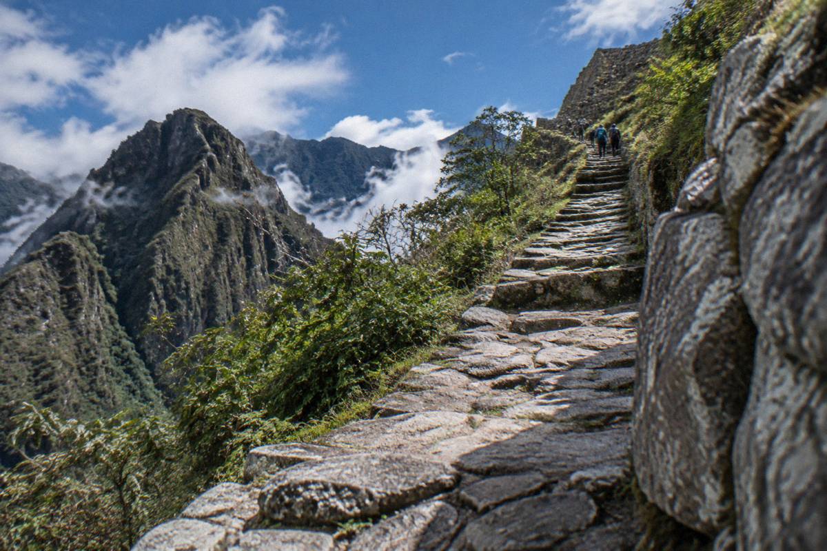 A narrow ancient stone-paved trail climbing through cloud forest with stone steps, a few tiny hikers far above, Machu Picchu