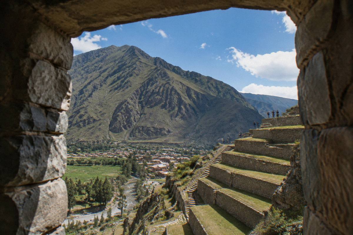 Stone terraces rising above the old Inca town of Ollantaytambo in the Sacred Valley, green fields and a river below, a few tiny visitors on a terrace