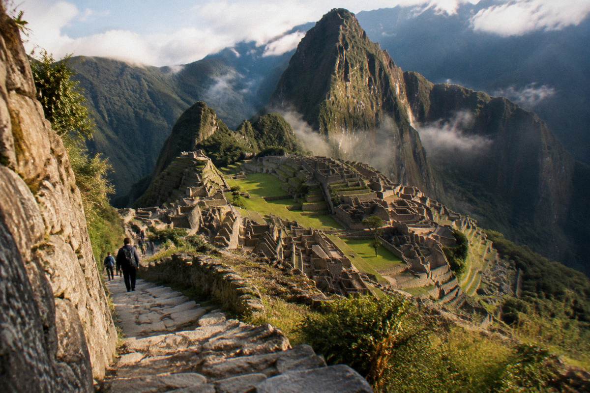 The terraced Inca citadel of Machu Picchu seen from a high ridge path, stone buildings among green peaks, thin cloud, a few tiny hikers on the path