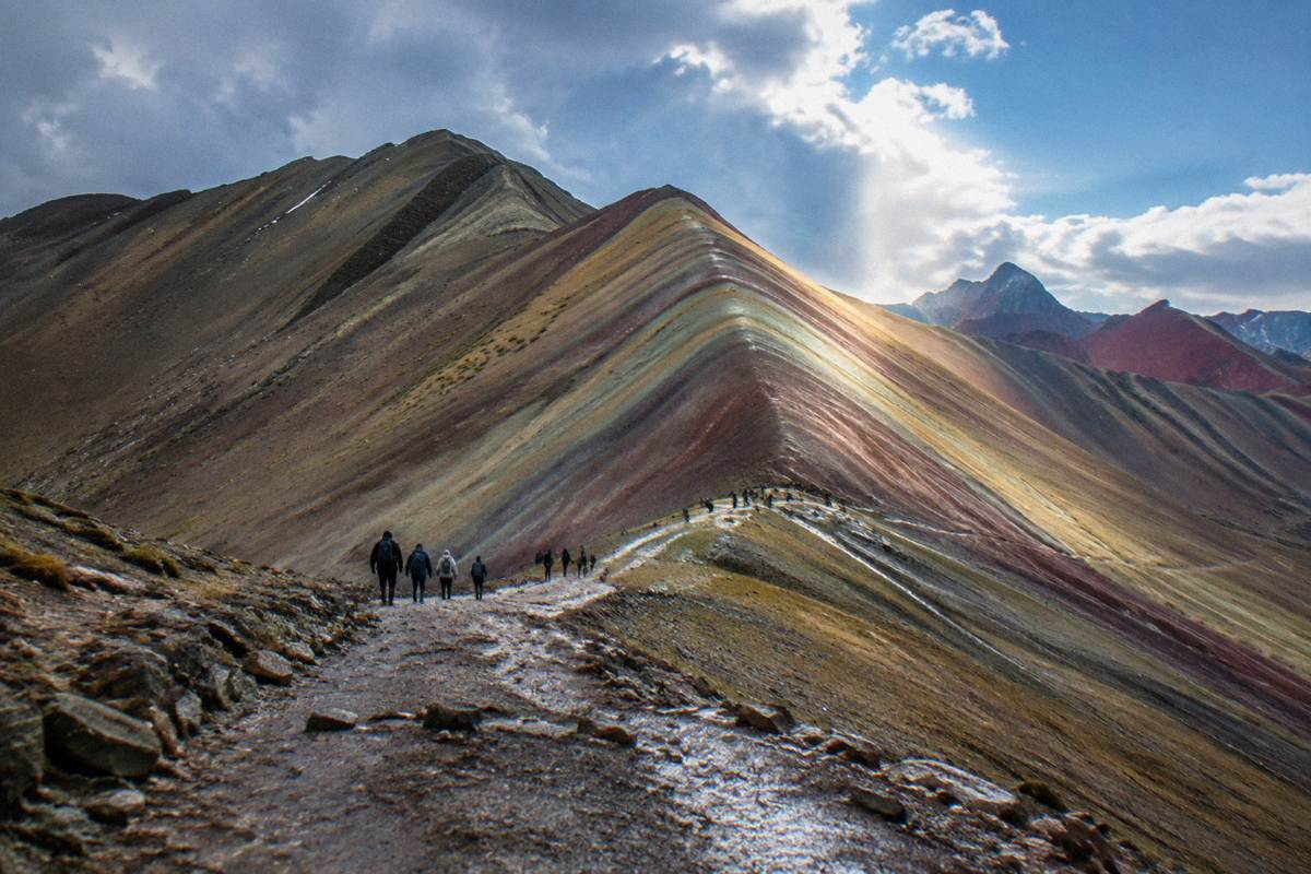 The striped red, ochre and turquoise ridge of Vinicunca under a cold blue sky, a line of tiny walkers on the path, Rainbow Mountain