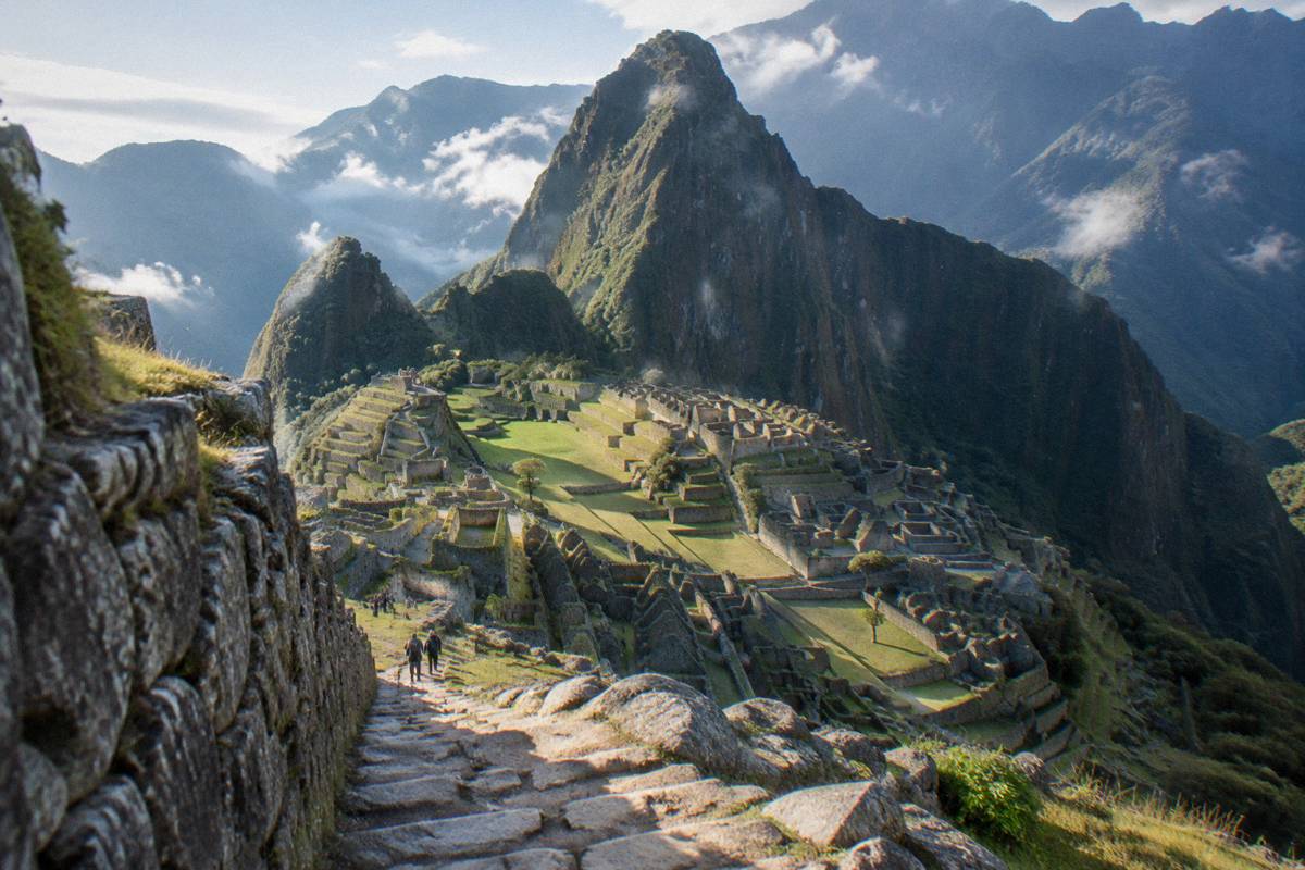 The Machu Picchu citadel seen from the classic upper viewpoint, stone terraces stepping down toward the steep pointed peak behind, thin cloud, tiny