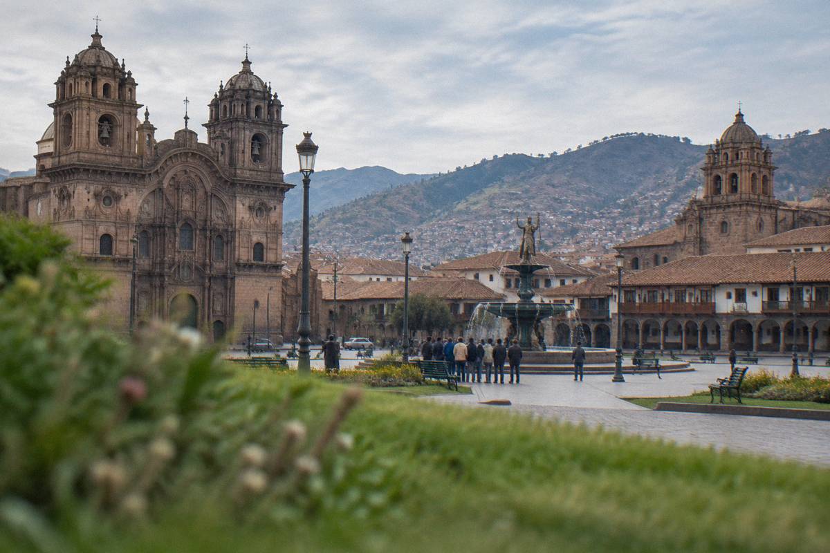 The Plaza de Armas in clear morning light, colonial arcades and stone church fronts, a small group gathered beside the fountain, seen from behind and far away, Plaza de Armas del Cusco