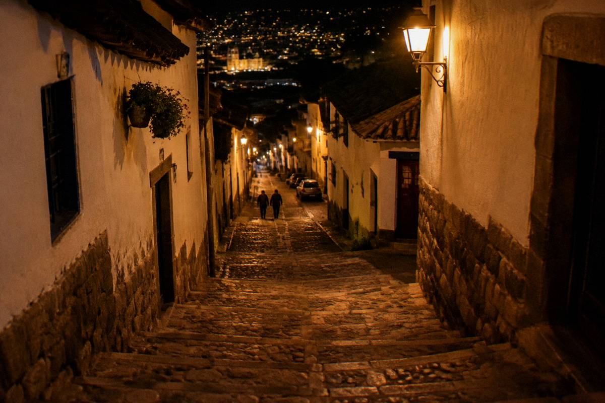 A steep stone stairway lane lit by warm lamps at night, whitewashed walls, a couple of figures walking uphill far away, San Blas