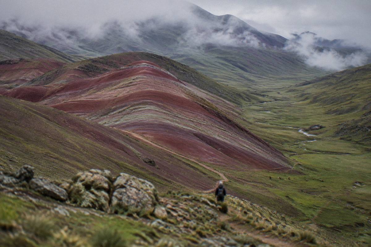 Banded red and mauve hills at Palccoyo rolling toward a green valley, a faint path, alpacas far off