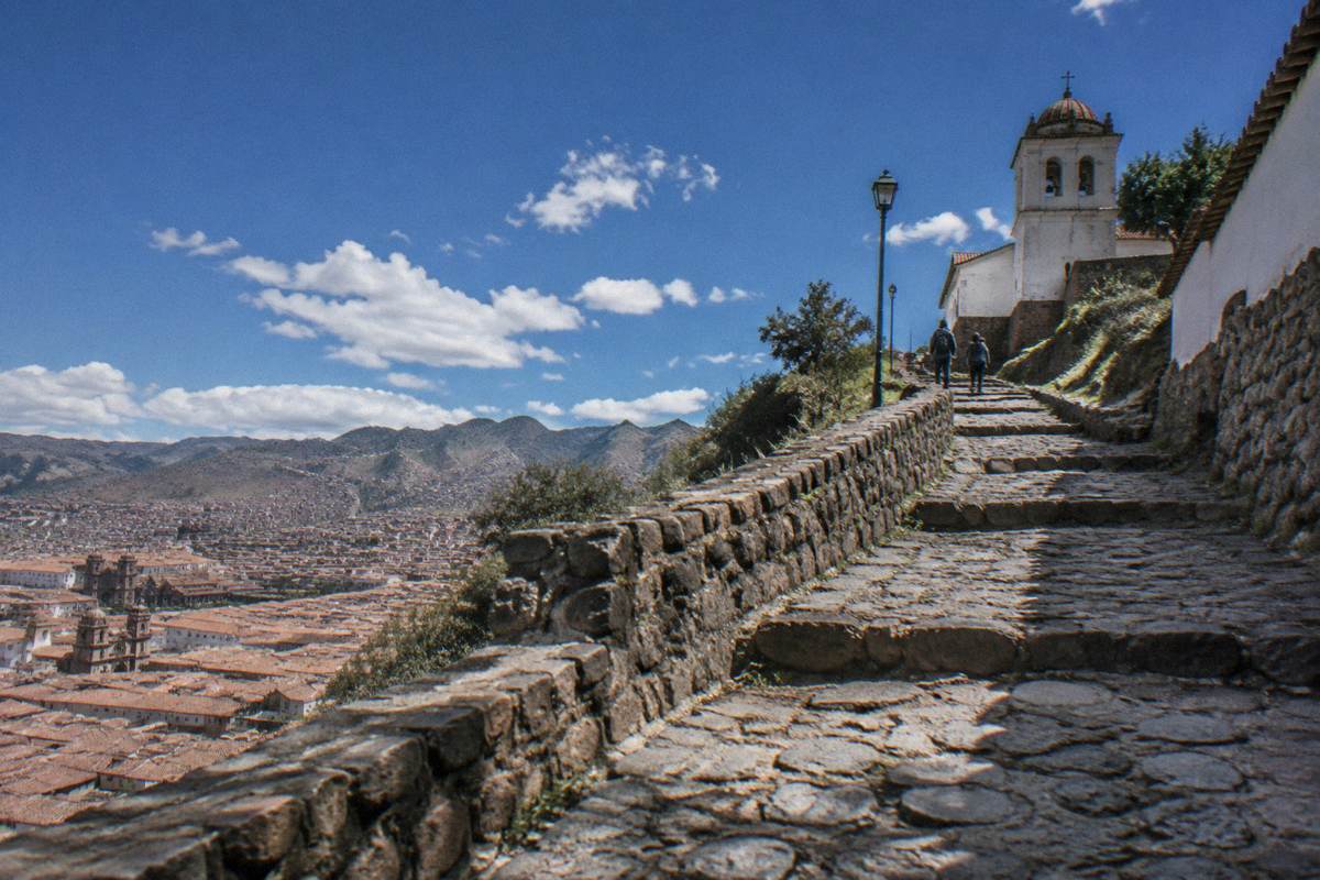 A hillside stone path climbing above Cusco's terracotta roofs toward a small whitewashed church, a pair of walkers far ahead, San Cristobal Church and Viewpoint
