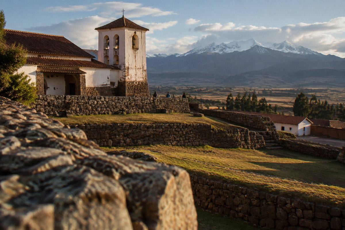 A stone-walled terrace below the colonial adobe church of Chinchero, snow peaks beyond a wide plateau