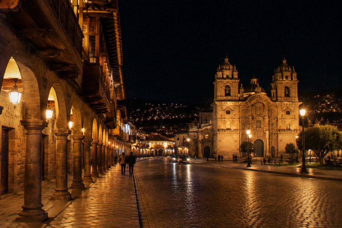 The arcaded Plaza de Armas at night, warm lamps along the arcades and a lit church front, a few small distant figures, Plaza de Armas del Cusco