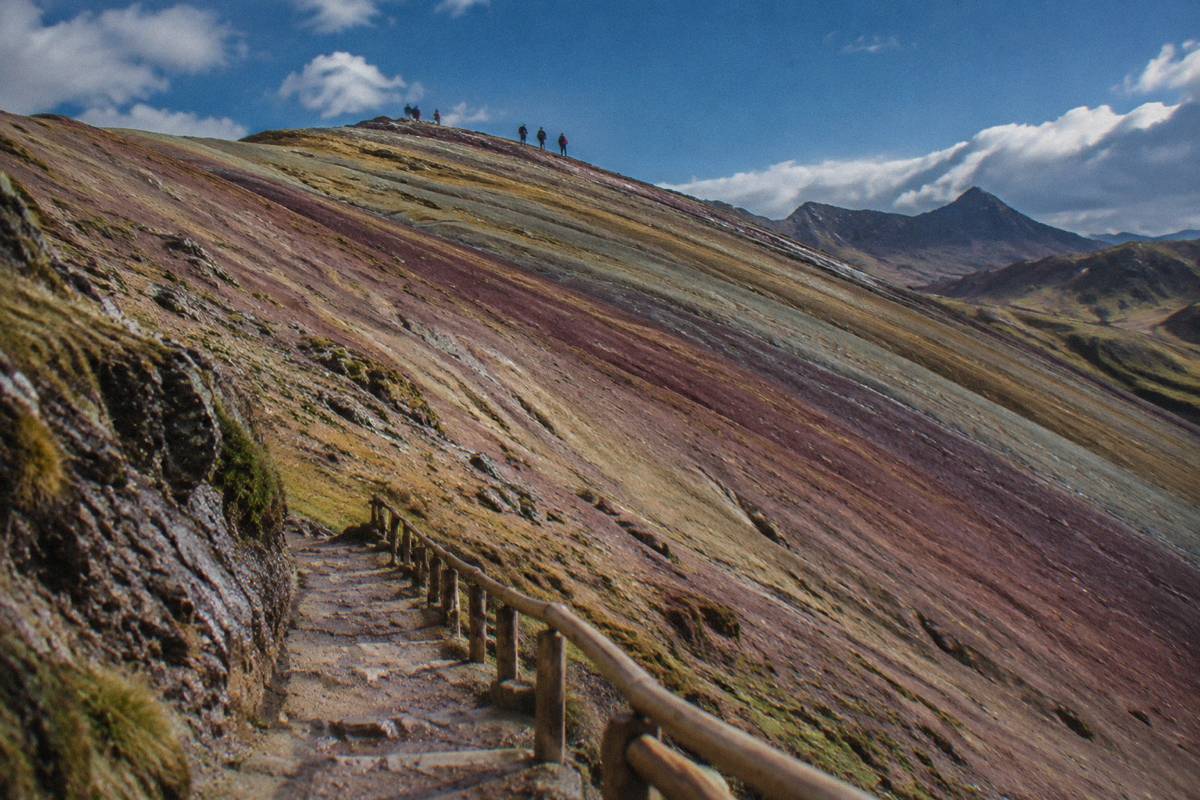 Layered violet, ochre and red stripes on a hill slope at Palccoyo seen close, a few distant hikers on the ridge under a deep blue sky