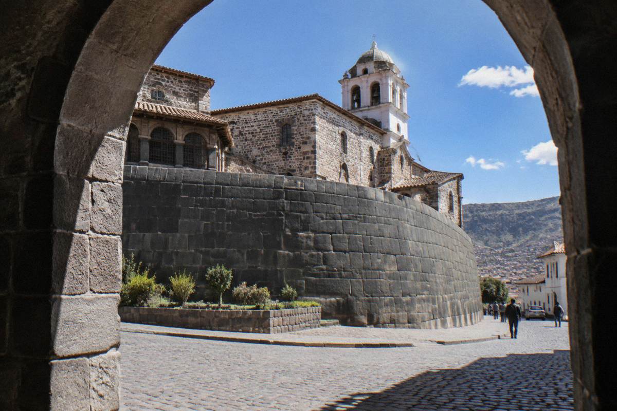 The curved Inca stone wall of Qorikancha with the colonial convent above it, a cobbled corner and a few passers-by far off
