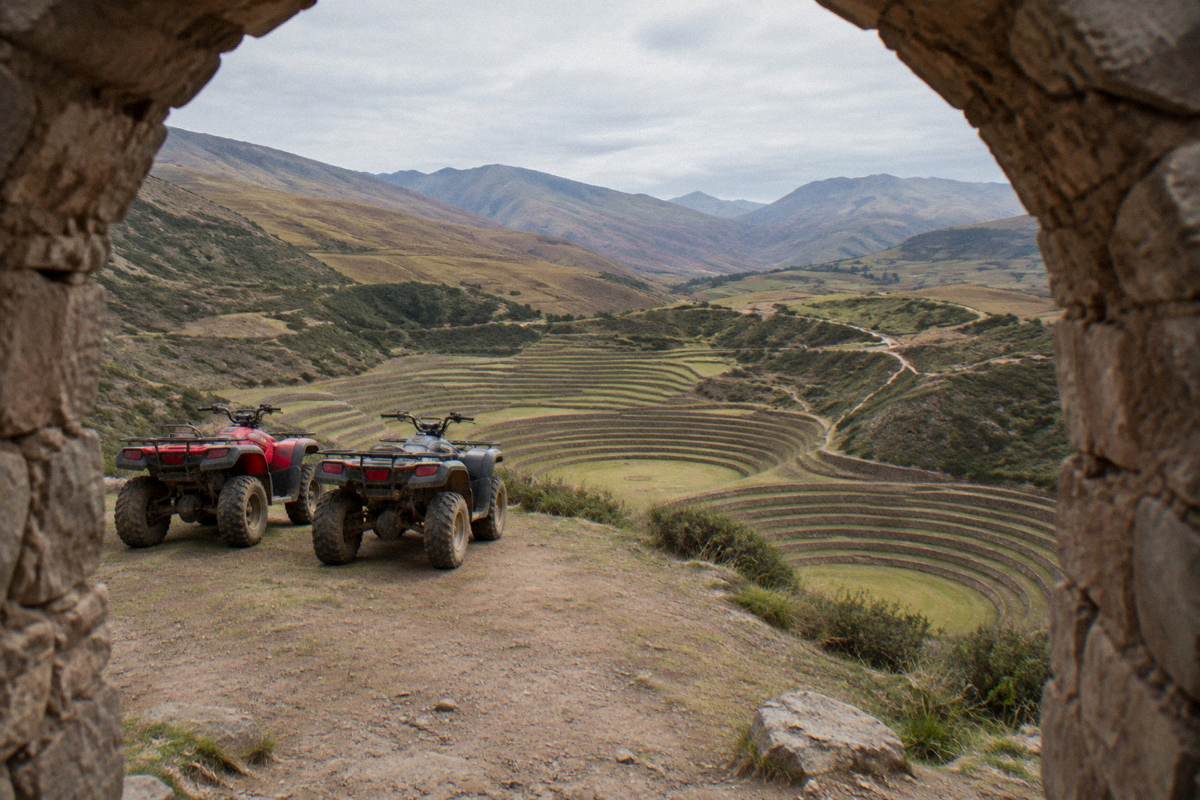 A pair of parked quad bikes on a dirt track on a high plateau overlooking the circular terraces of Moray, nobody on them