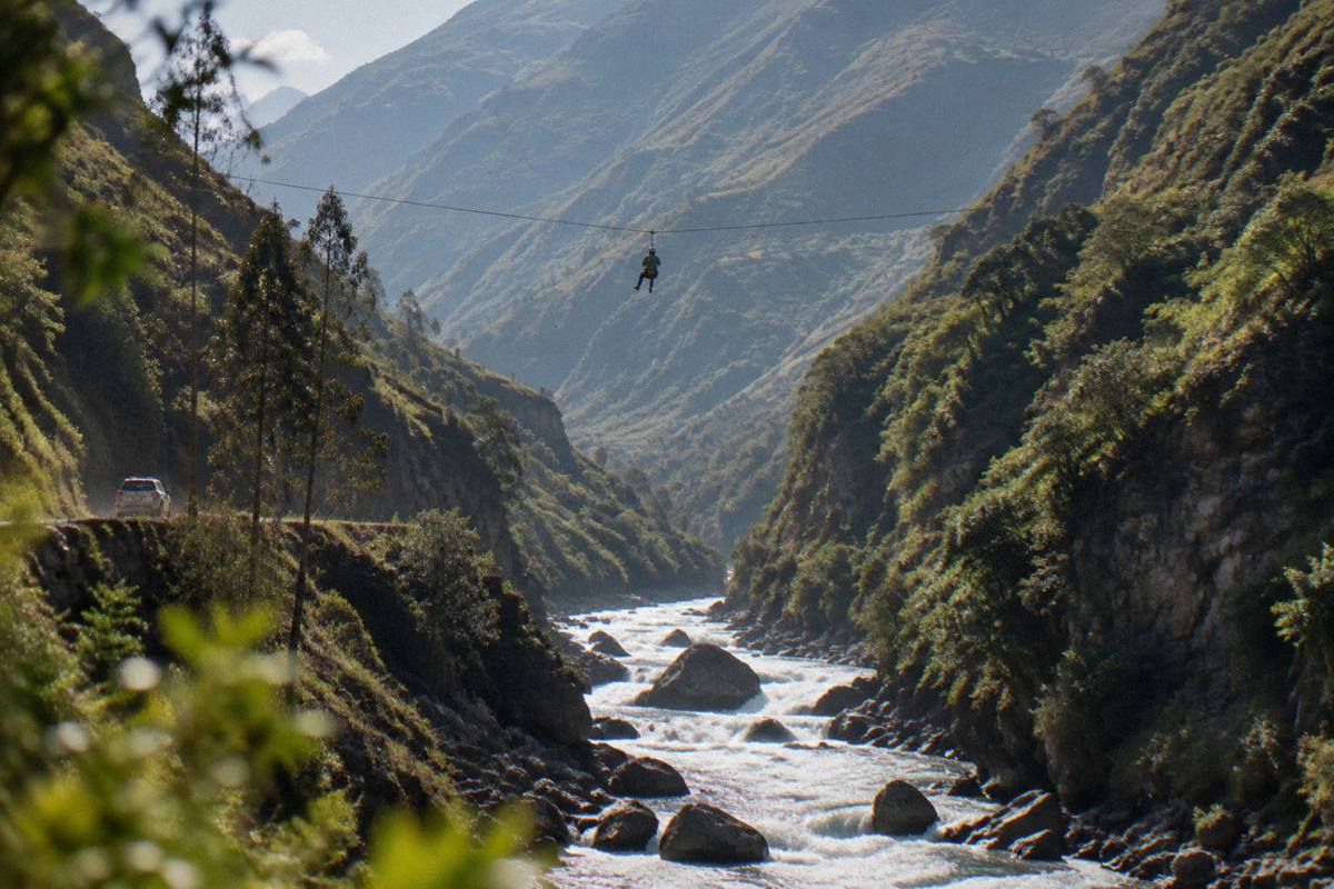 A fast river running over boulders under steep green slopes, a zip line cable crossing the gorge high above, Urubamba
