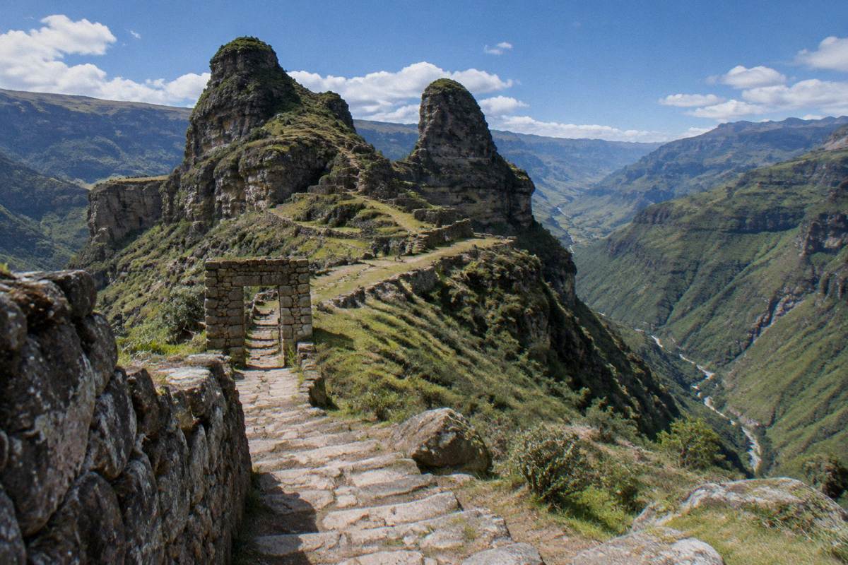 The horn-shaped stone summits and a ridge-top stone gateway of Waqrapukara over a deep green valley, a few tiny distant hikers