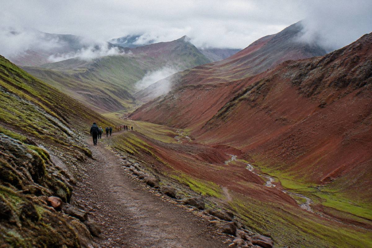 A deep red canyon valley with rust-red slopes and a trail near Vinicunca, a distant group of hikers