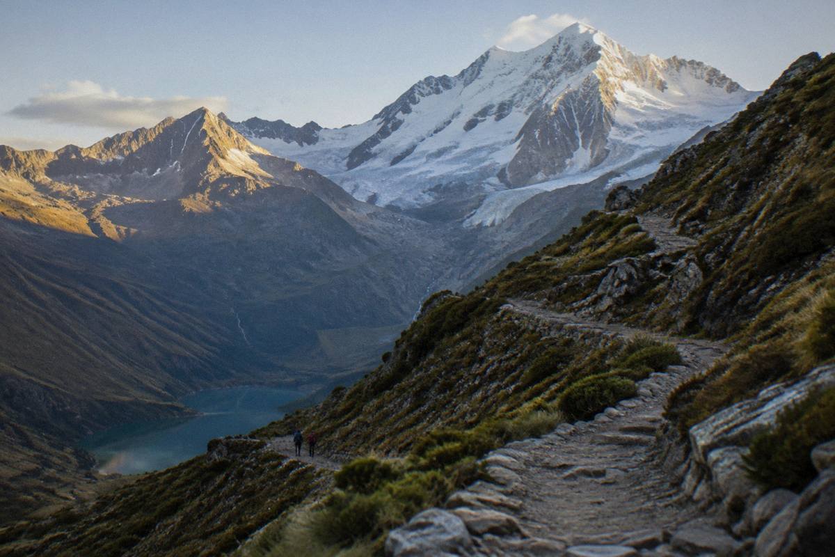 A rocky switchback trail climbing above a high valley toward a glacier peak in low morning light, two small walkers far below, Humantay Lake