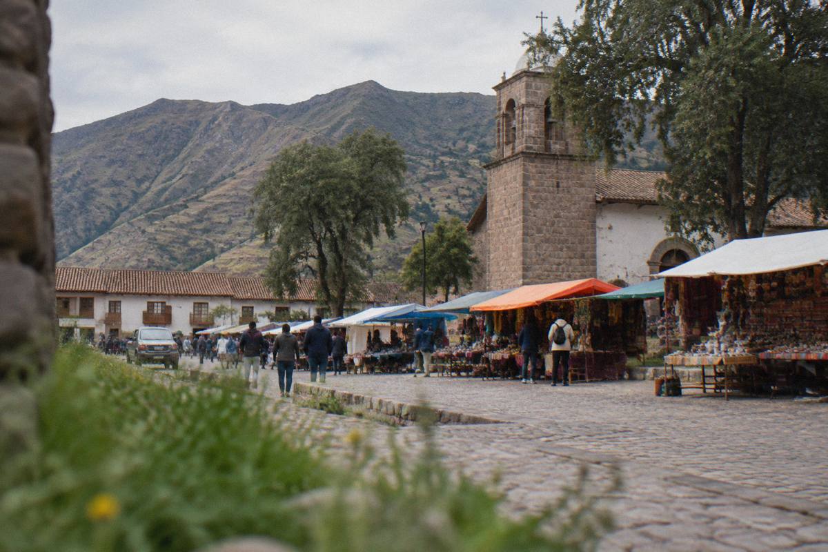 A lively Andean market town plaza with colourful stalls under awnings, a stone church and trees, shoppers small and from behind, Pisac town