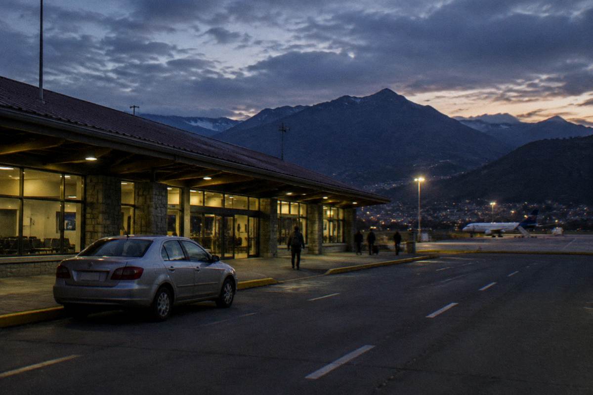A plain sedan parked at the kerb of a small airport terminal at dusk with mountains behind, Alejandro Velasco Astete International Airport