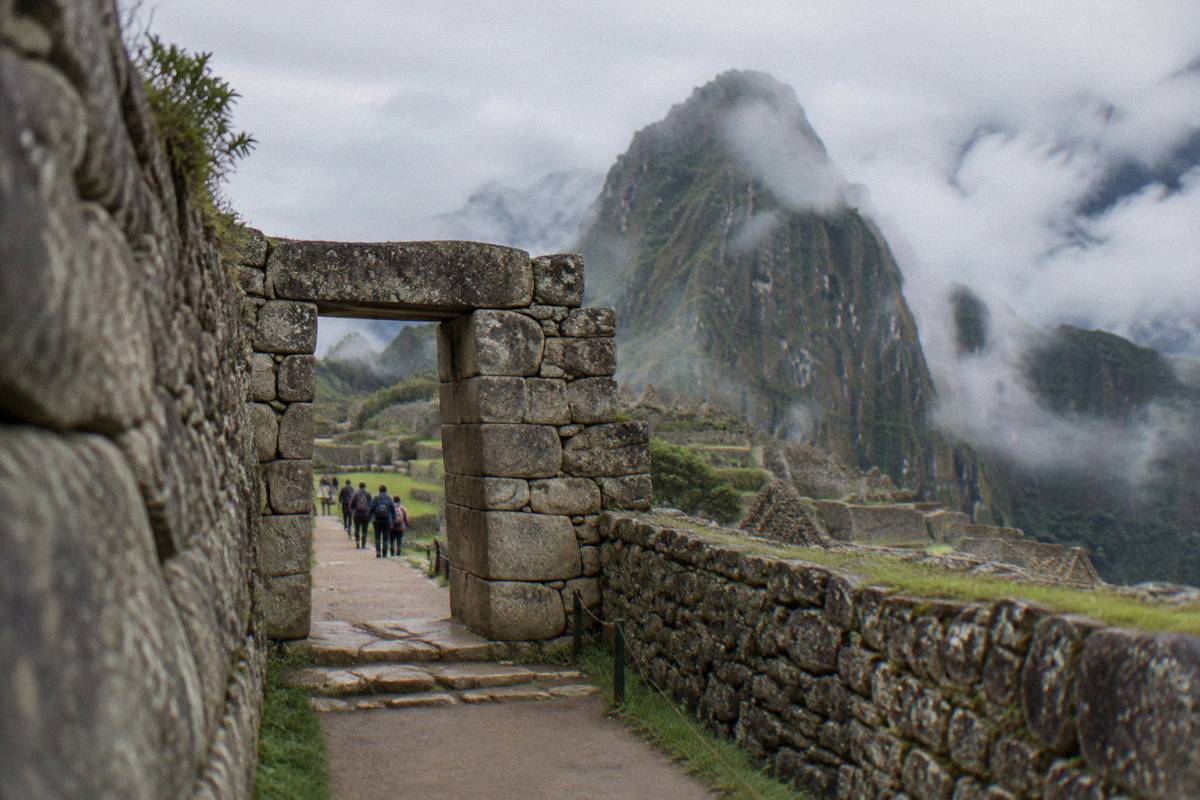A quiet stone gateway at the start of the path into the Machu Picchu ruins, steep green slopes and cloud behind, a few tiny visitors approaching