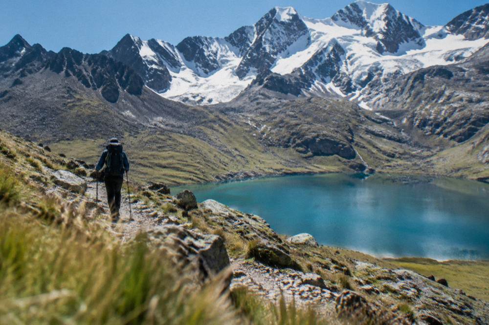 A trekker on a stony path above a blue-green lake with snow on the ridge