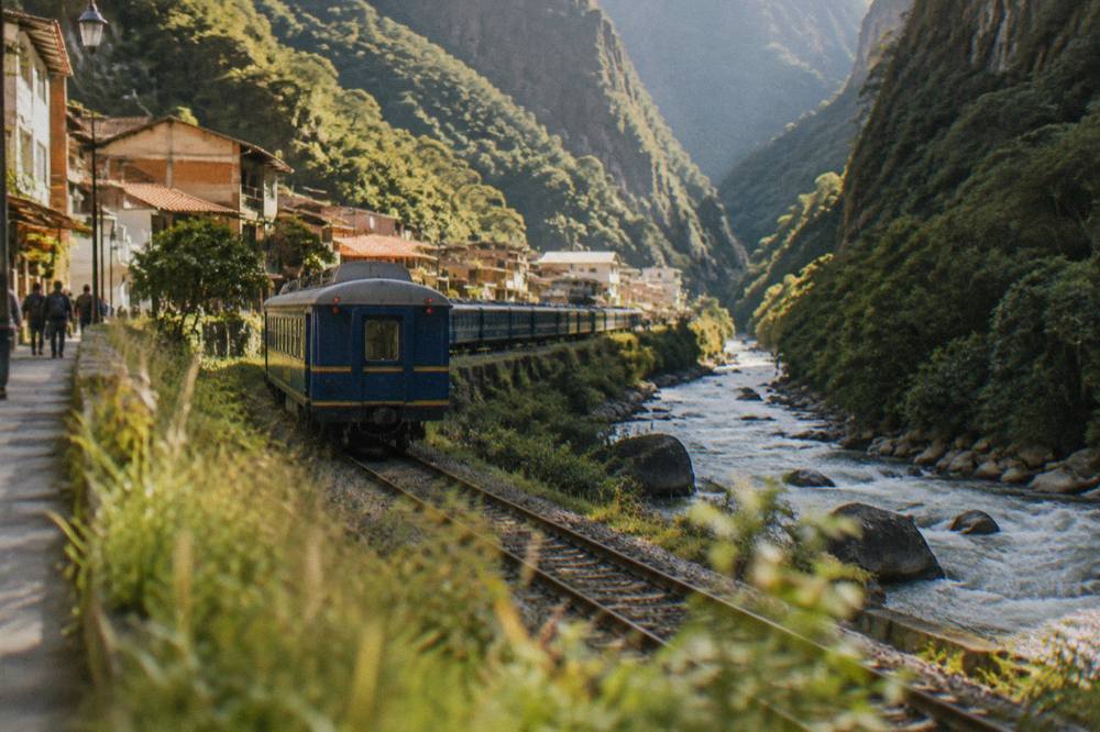 A passenger train on rails beside a river in a steep green valley