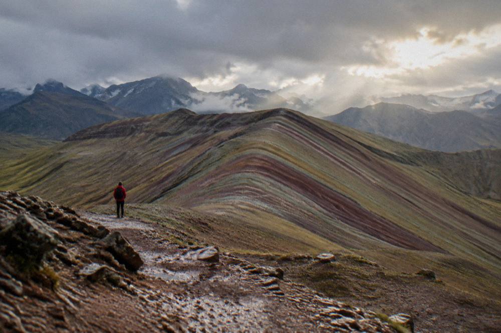 A hiker in a red jacket standing small against a vast striped mountain landscape
