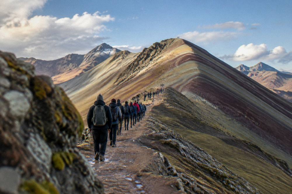 A long queue of walkers on a narrow ridge path under a bright blue sky