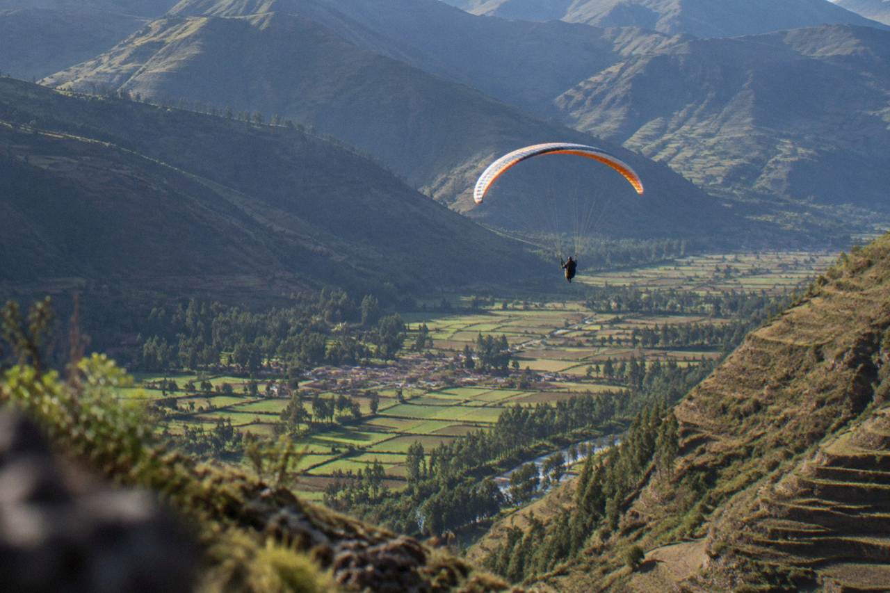 A paraglider flying above green Sacred Valley farmland and terraced hillsides in Peru
