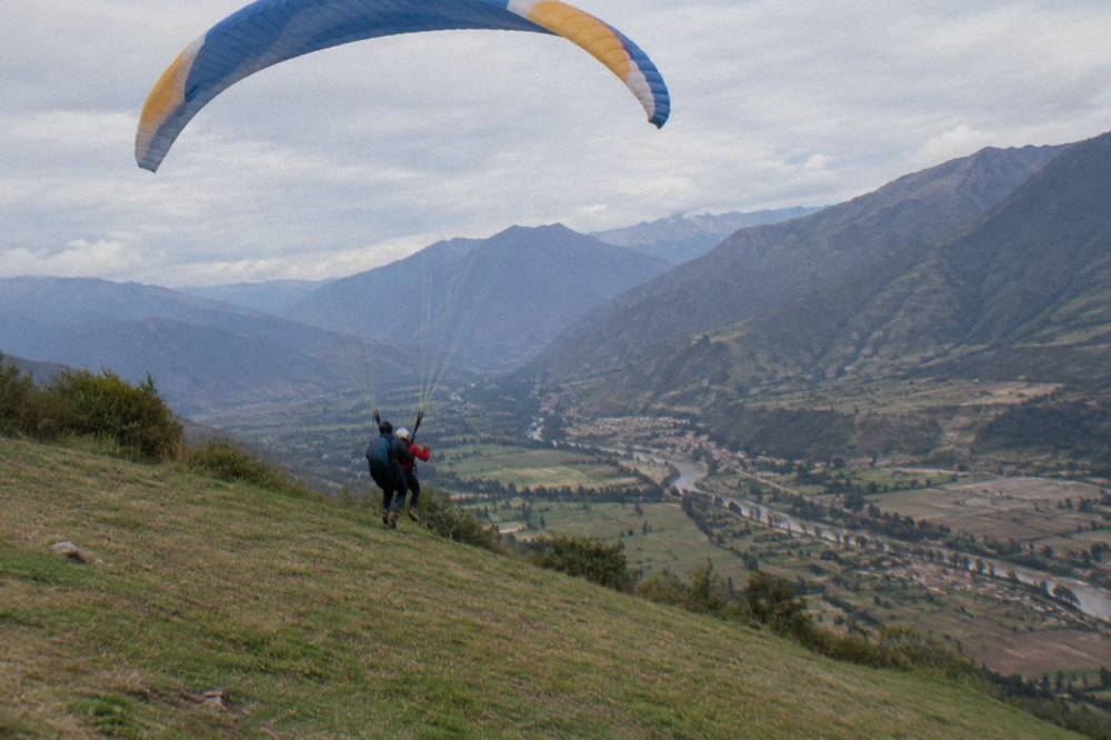 A tandem paraglider launching from a grassy slope above a wide Andean valley